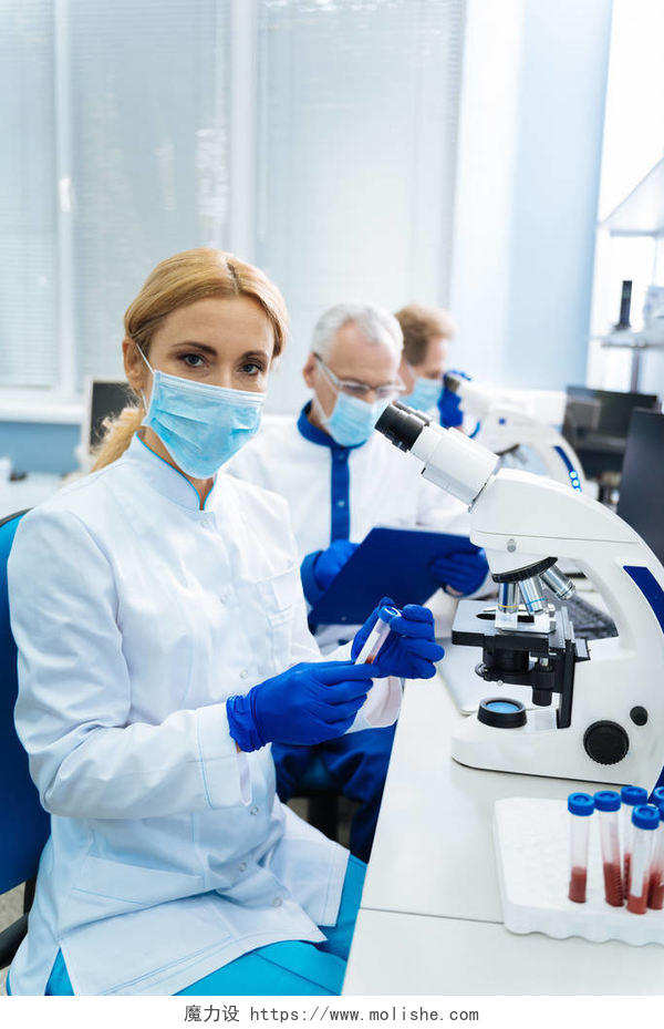 严肃的金发年轻研究员坐在显微镜和穿着制服和医疗口罩和持有试管